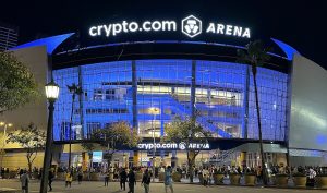 Nighttime view of the Crypto.com Arena in Los Angeles, lit in blue with a crowd gathered outside for an event.