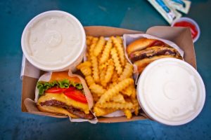 Overhead view of a takeout box with two cheeseburgers, crinkle-cut fries, and two covered soft drinks, with ketchup and condiments visible in the background.