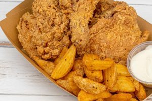 A cardboard container filled with crispy fried chicken, seasoned potato wedges, and a side of white dipping sauce on a white wooden surface.