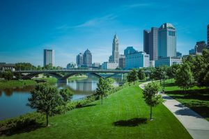 Columbus, Ohio skyline on a clear day with modern high-rise buildings, the Scioto River, and a green park with walking and biking paths in the foreground.
