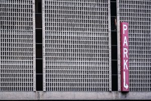 Exterior view of a parking garage with a grid-like concrete facade and a vertical red sign reading “PARK” with a white arrow pointing down.