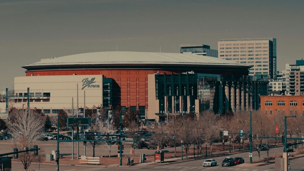 Exterior view of Ball Arena in Denver, Colorado, with downtown buildings in the background and light traffic in the foreground on a clear day.