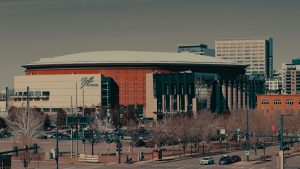 Exterior view of Ball Arena in Denver, Colorado, with downtown buildings in the background and light traffic in the foreground on a clear day.