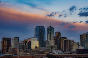 Denver skyline at sunset with modern high-rise buildings, including the Optiv tower, set against a dramatic sky with pink and orange clouds.