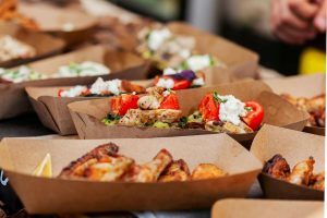 A variety of colorful gourmet dishes served in brown paper food containers, featuring grilled chicken, roasted vegetables, and dollops of cheese.