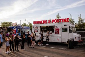 People line up at a white food truck labeled “Burgers & Poutine” during a food festival, with other colorful food trucks and trees in the background.