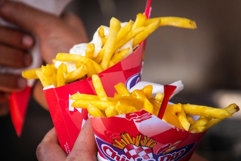 Close-up of two servings of French fries in red paper containers, one topped with melted cheese and the other with white dipping sauce, held by people at a food stand.