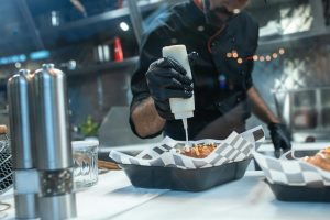 Man wearing black gloves and chef uniform drizzling white sauce over a hot dog in a food tray lined with checkered paper inside a modern kitchen