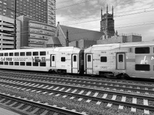 NJ Transit train cars at a station in Newark, New Jersey, with a church and tall buildings in the background, captured in black and white.