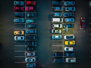 Overhead nighttime view of a parking lot filled with rows of parked cars, including a few brightly colored vehicles and a lit pay station near the center.