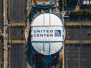 Overhead aerial view of the United Center in Chicago, with the arena roof showing the United Airlines logo and surrounding empty parking lots.