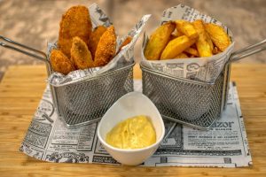 Crispy breaded potato wedges and thick-cut fries served in metal baskets lined with newspaper-style paper, with a side of creamy dipping sauce in a white bowl.
