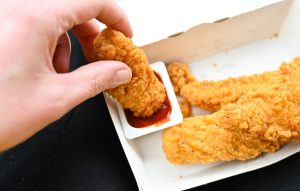Close-up of a hand dipping a crispy fried chicken tender into a container of red dipping sauce, with more chicken tenders in a white takeout box.