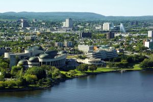 Aerial view of downtown Ottawa, Canada, featuring the curving architecture of the Canadian Museum of History near the Ottawa River, with residential and commercial buildings in the background and forested hills in the distance.