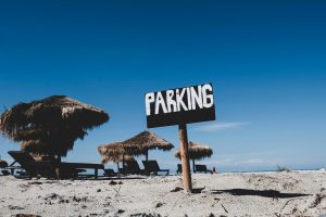 Weathered "PARKING" sign staked in sandy beach with straw umbrellas, lounge chairs, and a bright blue sky in the background.