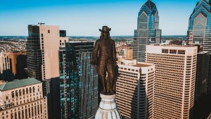 Aerial view of downtown Philadelphia featuring the statue of William Penn atop Philadelphia City Hall, surrounded by modern skyscrapers on a clear day.