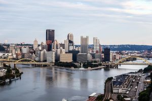 A wide view of downtown Pittsburgh, Pennsylvania, showing the city skyline with the U.S. Steel Tower and PPG Place, yellow bridges crossing the rivers, and the fountain at Point State Park where the Allegheny and Monongahela Rivers meet to form the Ohio River.