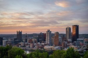 A scenic view of downtown Pittsburgh at sunset, featuring prominent skyscrapers including the PPG Place with its gothic-style spires, BNY Mellon Center, and the UPMC building reflecting golden light, with green trees in the foreground and soft clouds in the sky.