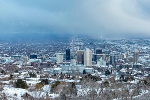 A panoramic winter view of downtown Salt Lake City, Utah, with snow-covered buildings and streets. The Utah State Capitol building is prominently featured in the foreground, with high-rise buildings and a snowstorm approaching in the distance.