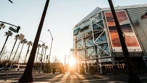 Exterior view of the SAP Center in San Jose, California, with the sun setting behind palm trees and street signs marking Santa Clara Street, creating a golden glow on the arena’s modern glass facade.