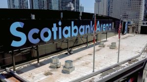 Rooftop view of Scotiabank Arena in Toronto, featuring large, bold light-blue letters spelling out the arena name. The foreground shows HVAC units and three flagpoles with Canadian and American flags. Skyscrapers of downtown Toronto rise in the background under a sunny sky.