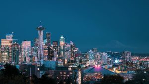 Seattle city skyline at night with the Space Needle illuminated in the foreground, skyscrapers lit up, and Mount Rainier faintly visible in the distance.