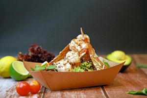 Gourmet shrimp taco served in a brown paper tray, garnished with lettuce, herbs, and a creamy sauce, surrounded by fresh ingredients including lime, avocado, and cherry tomatoes.