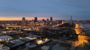 Aerial view of downtown St. Louis at dusk with city lights glowing, the Gateway Arch visible near the Mississippi River, and a vibrant sunset on the horizon.