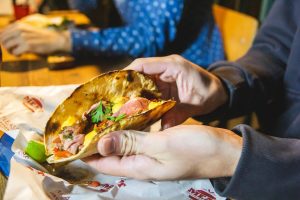 Close-up of a person holding a freshly made taco filled with grilled meat, melted cheese, pico de gallo, and cilantro, with a lime wedge and branded paper wrapping underneath on a table.