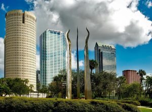 A view of downtown Tampa, Florida, featuring several modern high-rise buildings including the cylindrical Rivergate Tower and the striped Bank of America Plaza. In the foreground, a public art sculpture with four tall, curved metallic spires rises from a landscaped park area with lush green trees and shrubs under a partly cloudy sky.