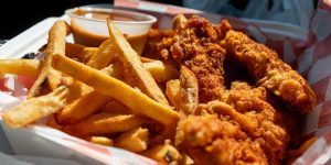A basket of crispy golden French fries and fried chicken tenders served in a red-and-white checkered paper-lined container with a cup of dipping sauce.