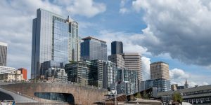 A view of downtown Seattle’s skyline featuring a mix of modern glass skyscrapers and older high-rise buildings, set against a partly cloudy sky. In the foreground, a curved wooden structure with people walking on top adds contrast to the tall buildings behind it.