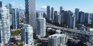 A view of downtown Vancouver with clusters of modern high-rise buildings, including a uniquely staggered tower, under a clear blue sky. A bridge with cars crosses in the foreground, connecting the dense cityscape.