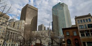A cityscape view of downtown Winnipeg, featuring tall modern office towers with reflective windows alongside older historic brick and stone buildings in the foreground. Leafless trees line the street under a partly cloudy sky.